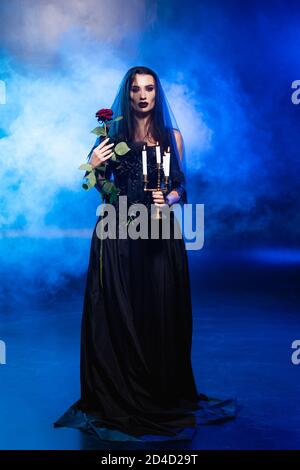 Braut in schwarzem Kleid und Schleier hält Rose und brennende Kerzen auf blau mit Rauch, halloween-Konzept Stockfoto