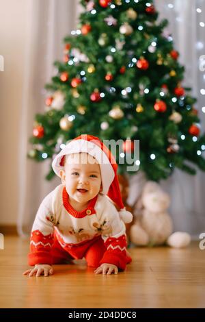 Baby in einem gestrickten Rentier Kostüm und Weihnachtsmann Hut ist Kriechen vor einem Weihnachtsbaum Stockfoto