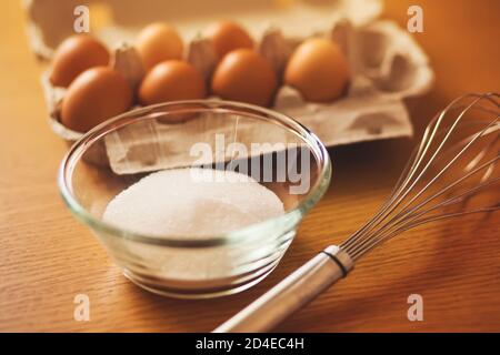 Auf einem Holztisch sind die Zutaten: Hühnereier in einer Packung und Zucker in einer Glasschüssel, und daneben ist ein Schneebesen, um alles zu mischen. Kochen Proce Stockfoto
