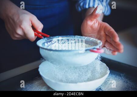 Ein Koch in einer blauen Schürze und einem gestreiften Hemd hält ein Sieb mit rotem Griff und verwendet es, um das krümelige Mehl in eine weiße Schüssel zu sieben. Hausmannskost. Stockfoto