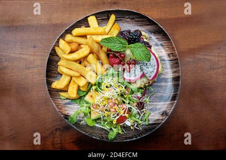 Pommes frites, gemischter Salat und ein mit Früchten überzogener Schnitzel auf einem Teller und auf einem Holztisch, Hochwinkelansicht von oben, ausgewählter Fokus Stockfoto