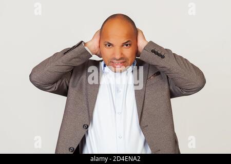 Verärgert Mann afroamerikanischen Geschäftsmann in Jacke, bedeckt seine Ohren mit Händen, hören lästige Geräusche, er hat Kopfschmerzen, posiert für Kamera und bedeckt seine Ohren durch Lärm. Stockfoto