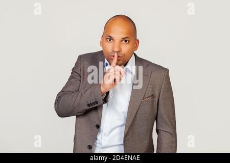 Ein junger afroamerikanischer Geschäftsmann in einer Jacke, der seinen Zeigefinger auf die Lippen drückt, bittet niemanden eine Geschichte zu erzählen, seinen Mund zu schließen und im falschen Moment nicht mehr zu scherzen. zeichen des Schweigens Stockfoto