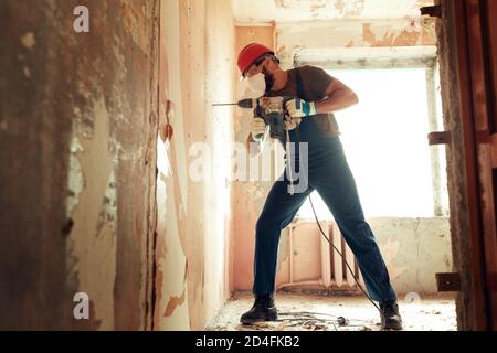 baumeister mit Perforator bohrt Löcher in Betonwand der Baumeister Ist in Schutzanzug und Helm gekleidet Stockfoto
