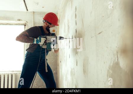 baumeister mit Perforator bohrt Löcher in Betonwand der Baumeister Ist in Schutzanzug und Helm gekleidet Stockfoto