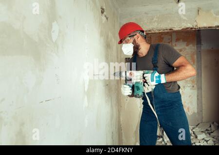 baumeister mit Perforator bohrt Löcher in Betonwand der Baumeister Ist in Schutzanzug und Helm gekleidet Stockfoto