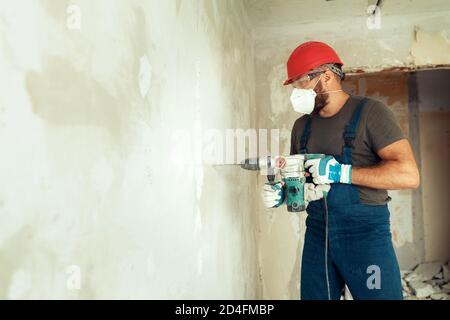 baumeister mit Perforator bohrt Löcher in Betonwand der Baumeister Ist in Schutzanzug und Helm gekleidet Stockfoto