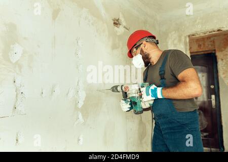 baumeister mit Perforator bohrt Löcher in Betonwand der Baumeister Ist in Schutzanzug und Helm gekleidet Stockfoto