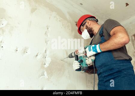 baumeister mit Perforator bohrt Löcher in Betonwand der Baumeister Ist in Schutzanzug und Helm gekleidet Stockfoto