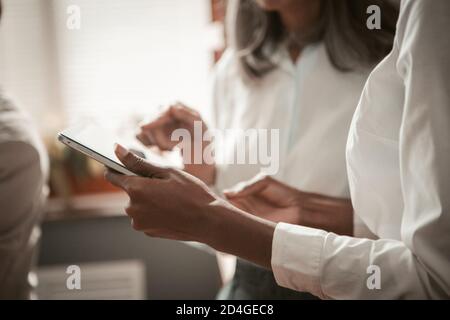 Geschäftsfrauen, die im Büro ein digitales Tablet nutzen. Nahaufnahme der weiblichen Hände. Afroamerikanische und asiatische Frauen tragen weiße Hemden, die von Sonnenlicht beleuchtet werden Stockfoto