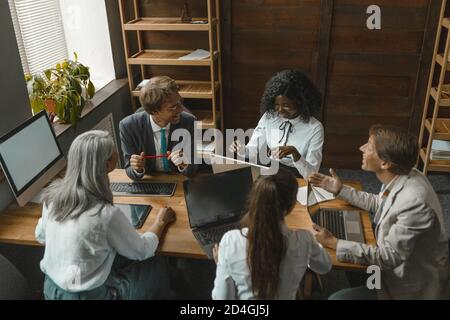 Teamarbeit in einer vielfältigen Gruppe. Fröhliche Büroangestellte lachen miteinander, während sie zusammen am Tisch sitzen und Computer darauf haben. Weitwinkelansicht. Getöntes Bild Stockfoto