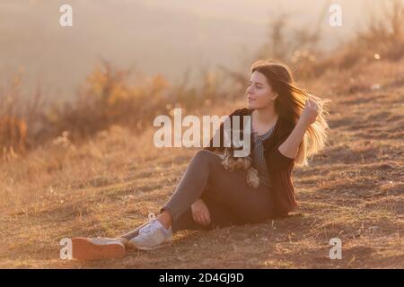 Schöne junge Mädchen mit langen roten Haaren Entwicklung im Wind umarmt einen schwarzen Schnauzer Hund. Sie schaut auf die Kamera und lächelt im Sonnenuntergang. Tierpflege Stockfoto