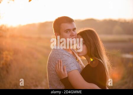 Ein junger Mann küsst, umarmt eine schöne lächelnde blonde Frau mit langen Haaren. Nahaufnahme eines Paares, das sich vor dem Hintergrund des Herbstes umarmt Stockfoto