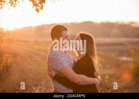 Ein junger Mann küsst, umarmt eine schöne lächelnde blonde Frau mit langen Haaren. Nahaufnahme eines Paares, das sich vor dem Hintergrund des Herbstes umarmt Stockfoto