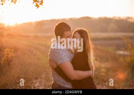 Ein junger Mann küsst, umarmt eine schöne lächelnde blonde Frau mit langen Haaren. Nahaufnahme eines Paares, das sich vor dem Hintergrund des Herbstes umarmt Stockfoto