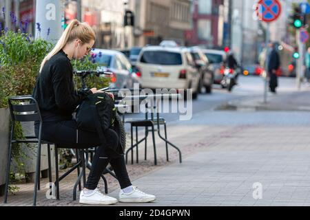 Riga, Lettland - 8. Oktober 2020: Eine junge Frau mit Sonnenbrille sitzt an einem Tisch auf dem Bürgersteig auf der Seite der Straße und trinkt Kaffee Stockfoto