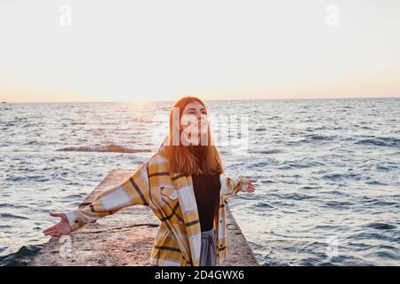 Glückliche, freie junge Frau, die bei Sonnenaufgang am Meer mit ausgestreckten Armen steht. Warmes Licht. Freiheit und Wohlfühlen Konzept. Stockfoto