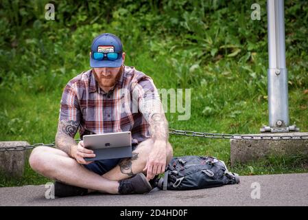 Junger Mann sitzt mit gekreuzten Beinen am Straßenrand mit seinem Apple iPad, im Schatten an einem heißen Sommertag. Schloss Neuschwanstein, Deutschland. Stockfoto