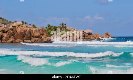 Charakteristisch schöne Granitfelsen mit Kokospalmen dazwischen und brechenden Wellen im türkisfarbenen Wasser am Strand Petite Anse, La Digue. Stockfoto