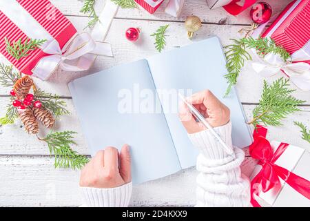 Weibliche Handschrift in Notizblock mit weihnachtsdekoration und Geschenkschachteln auf weißem Holztisch. Weihnachtswunsch, weihnachtsbrief, To-Do-Liste Konzept, Stockfoto