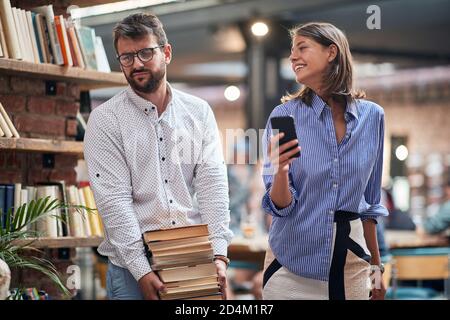 Ein junger Mann, der seiner Freundin wegen schwerer Bücher widerwillig ist Sie wählten in der Bibliothek Stockfoto