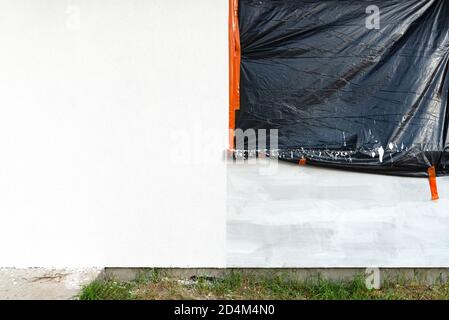 Eine neu aufgetragene Schicht aus weißem Silikonputz an der Hauswand, das Fenster ist mit schwarzer Folie gesichert. Stockfoto