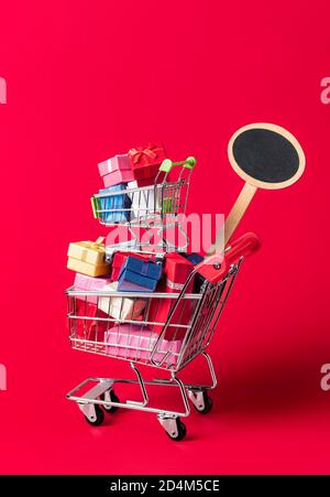 Weihnachts-Shopping-Konzept mit einem Warenkorb voller Geschenke und ein leeres Schild für Verkauf und Rabatt. Warenkorb mit Geschenken isoliert auf rotem Hintergrund Stockfoto