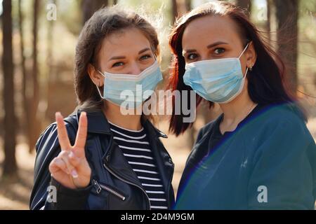 Zwei junge Frauen in medizinischen Masken betrachten die Kamera in Holz. Nahaufnahme von Frauen schützen sich vor Krankheiten auf dem Spaziergang. Konzept der Bedrohung von Stockfoto