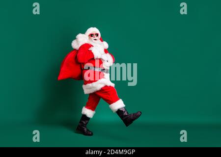 In voller Länge Profil Foto von im Ruhestand Opa weißen Bart gehen Mit Sack Geschenke Kinder tragen roten weihnachtsmann Kostüm Mantel Handschuhe Sonnenschutz Stockfoto