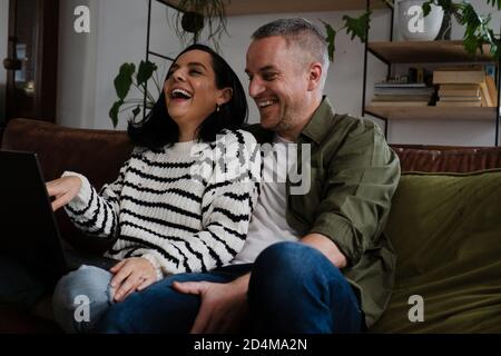 Glückliches Paar, das lachte, während es sich Videos auf einem Laptop ansah, der auf einem bequemen Sofa in der Lounge saß. Stockfoto