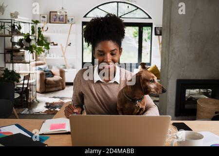 Lächelnde Studentin auf Videoanruf, die Freunde von der Universität aufholt, mit dem Welpen sitzend, der in der modernen Lounge studiert. Stockfoto