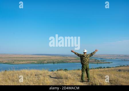 Glücklicher Mann in einem Hut hebt seine Hände auf den Berg. Vor dem Hintergrund von Fluss und blauem Himmel. Kopierbereich, Softfokus. Freiheit und Reisen Stockfoto