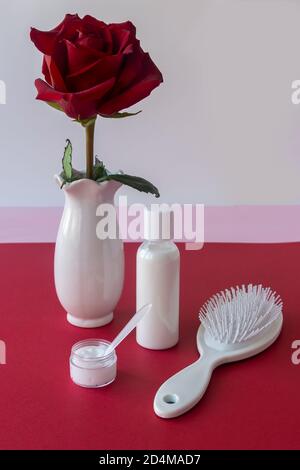 Weißer Haarkamm, unbeschriftete Shampoo-Flasche, Haarpflegeprodukte und rote Rose in weißer Vase auf dem Tisch. Konzeption von Naturkosmetik, Spa-Behandlungen Stockfoto