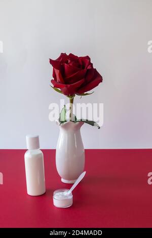 Gesichts- und Körperpflegeprodukte in weißer Flasche und Dose ohne Marke auf dem Tisch. Rote Rose in weißer Vase, Farbkontrast. Das Konzept der Kosmetologie, Spa Stockfoto