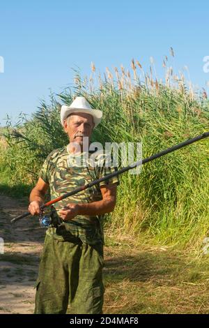 Senior Fisherman fängt Fische am Flussufer in Schilf. Angeln im Ruhestand im Sommer. Helles Sonnenlicht, weicher Fokus. Stockfoto