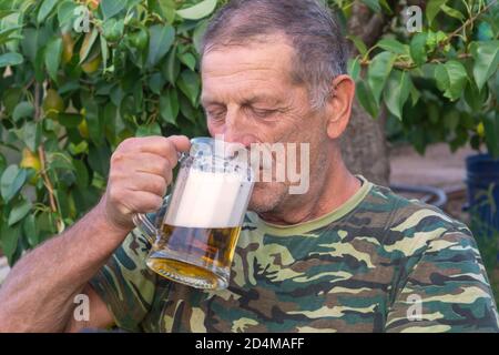 Großvater trinkt an heißen Sommertagen im Freien schaumig kaltes Bier aus einem Becher. Entspannung und Alkoholkonzept, selektiver Fokus, Nahaufnahme. Stockfoto