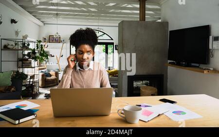 Besorgte Geschäftsfrau mit Ohrhörern, die bei einem Videoanruf neben der Papierarbeit chattet und im schicken Wohnzimmer einen heißen Kaffee schneidet. Stockfoto