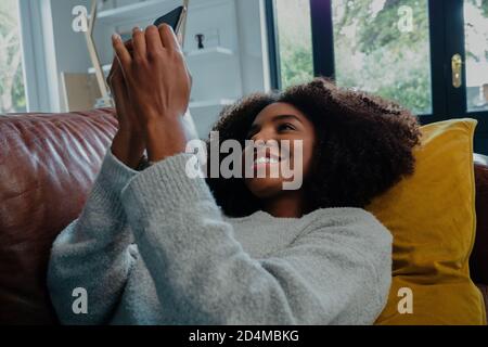 Gemischte Rasse Frau mit afro glücklich SMS auf Smartphone liegend auf Ledersofa in kausalen Lounge. Stockfoto