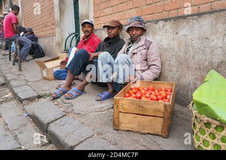 Antananarivo, Madagaskar - 24. April 2019: Drei unbekannte madagassische Männer sitzen auf dem Bürgersteig und verkaufen Tomaten. Essen wird in der Regel auf den Straßen in Madaga verkauft Stockfoto