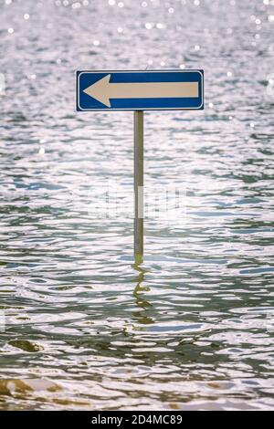 Plakatwand-Straßenpfeil nach links teilweise von Hochwasser überflutet. Mitten in einem See liegt ein Straßenschild. Stockfoto