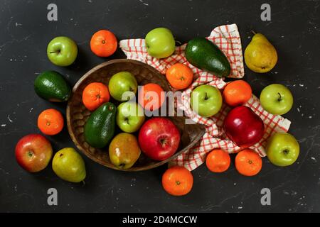 Äpfel, Birnen, Orangen und Avocado in geschnitzter Holzschale auf schwarzem marmorähnlichen Brett, Blick von oben Stockfoto