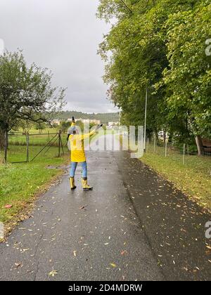 Fröhliche Frau in gelbem Regenmantel und Regenstiefeln mit jaguar Muster zu Fuß an regnerischen Tag. Rückansicht der Person. Stockfoto