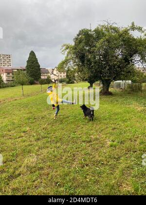 Fröhliche Frau in gelbem Regenmantel und Regenstiefeln mit jaguar-Muster, die am Regentag im Herbst mit Hund spielt. Stockfoto