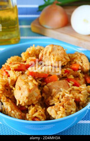 Reis mit Huhn und Gemüse. Blauer Teller. Nationalgericht Osten. Im Hintergrund ein Schneidebrett mit Zwiebeln und geschnittenen Karotten. Stockfoto