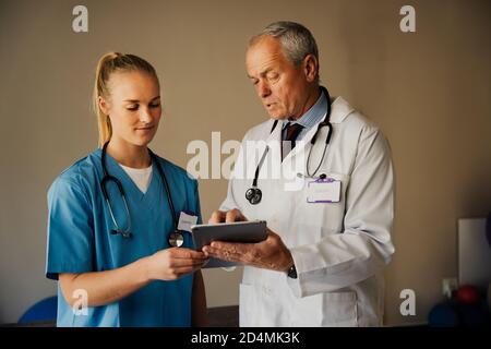 Männlich Arzt Lehre weibliche Praktikantin in Scrubs, während zeigt Anweisungen auf digitale Tablette in organisierten Ärzten Zimmer Stockfoto