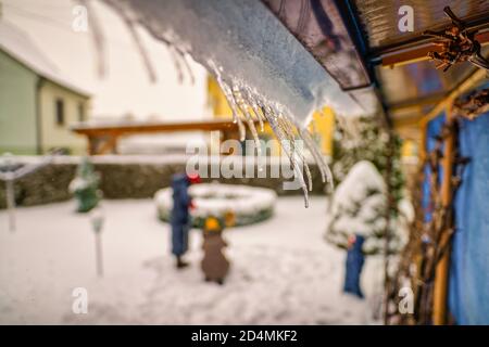 Eiszapfen hängen vom Hausdach in einem Garten mit einer verschwommenen Familie im Hintergrund - typische Winterlandschaft. Stockfoto