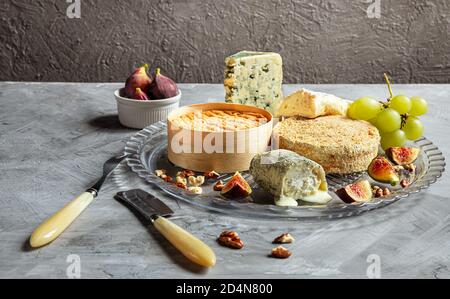Auswahl an französischem Käse - Camembert, roquefort, Brie, Ziegenkäse und Epoisse mit Trauben, Feigen und Nüssen auf grauem Hintergrund Stockfoto