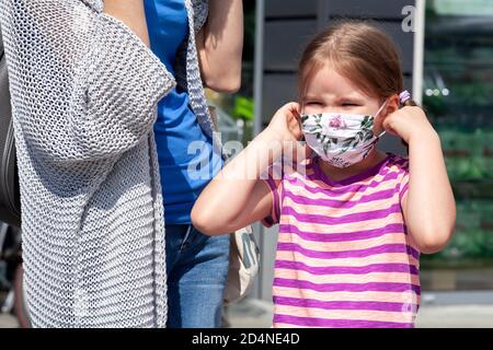 Mutter und Tochter zusammen, kleines Mädchen, das an- oder auszieht schützende antivirale medizinische Gesichtsmaske. Familieneinkäufe während der Pandemie Stockfoto