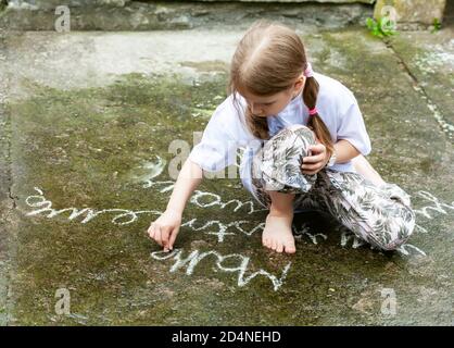 Kleines junges Mädchen, das einfache Wörter auf Beton mit weißer Kreide schreibt. Kinderzeichnung mit Kreide auf dem Boden im Freien. Kunsttherapie-Konzept. Schulkind Stockfoto