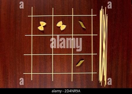 Italienische Pasta. TIC Tac Toe oder XS und OS Spiel aus rohen Spaghetti, Farfalle und Penne gemacht. Trocknen Sie Makkaroni und Capellini auf einem braunen Holztisch. Stockfoto